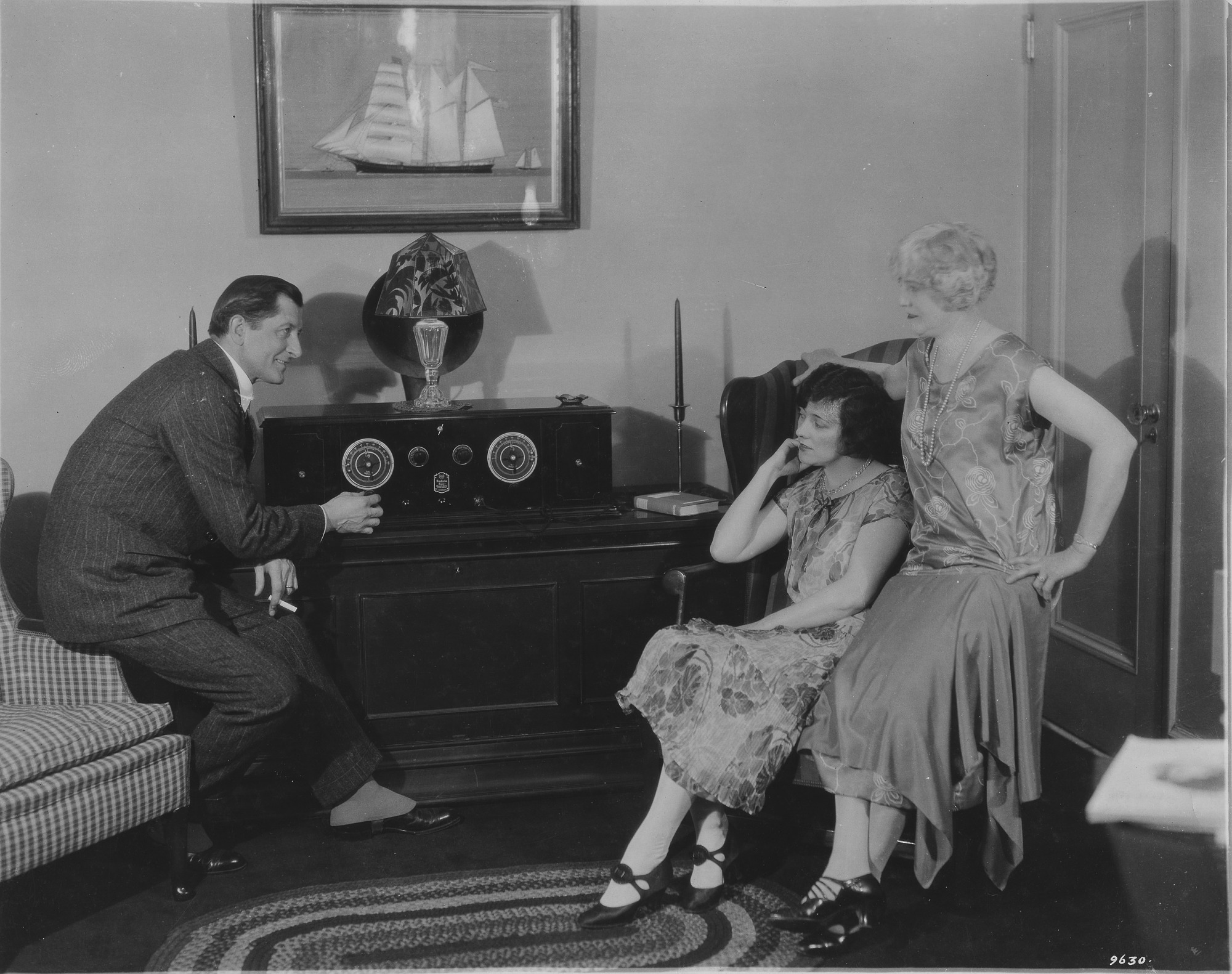 Personer i 1920-talsklädsel runt en antik radio i ett vardagsrum